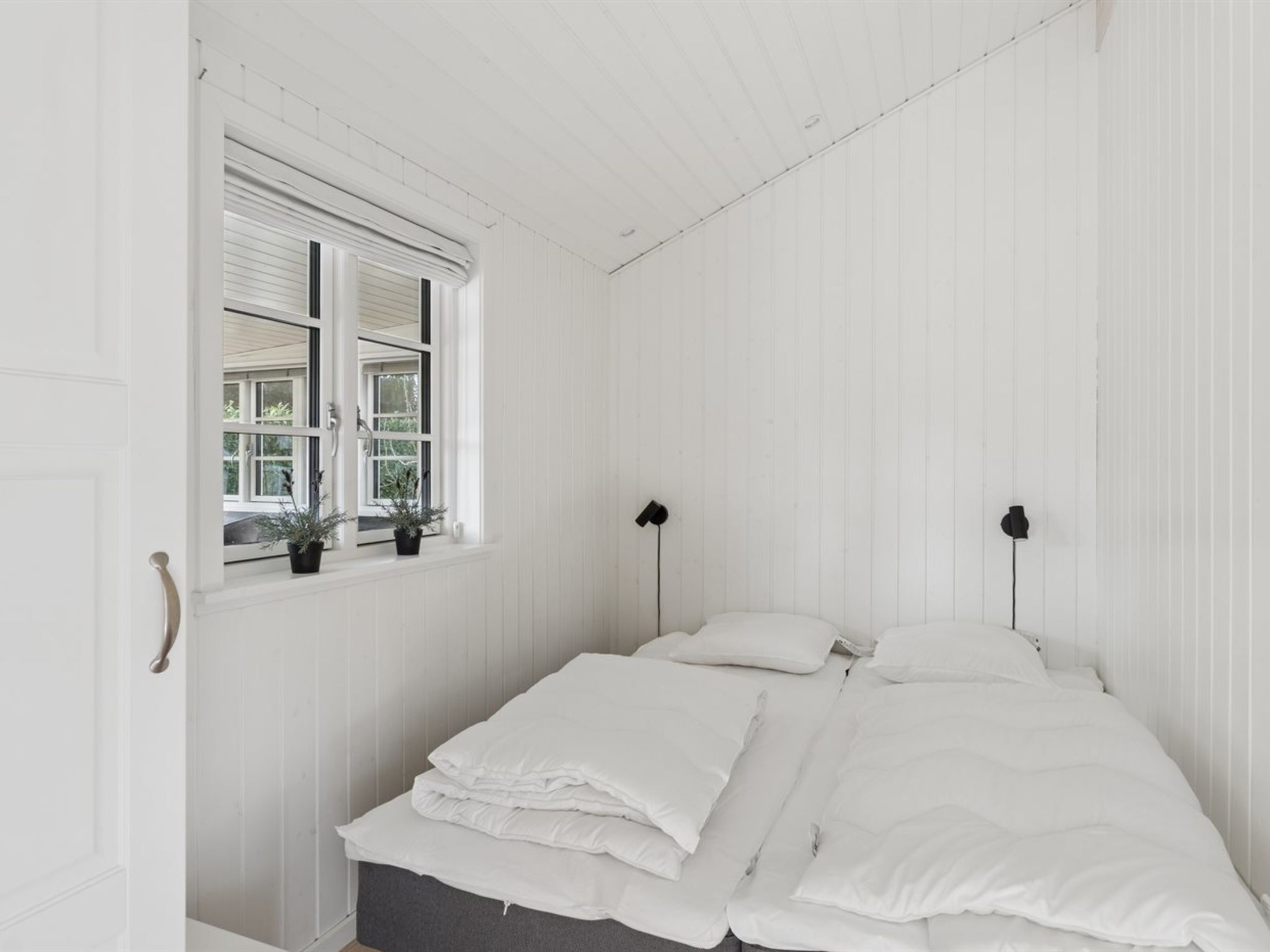 A white room with a bed, two bedside lamps, a window with plants, and a door.