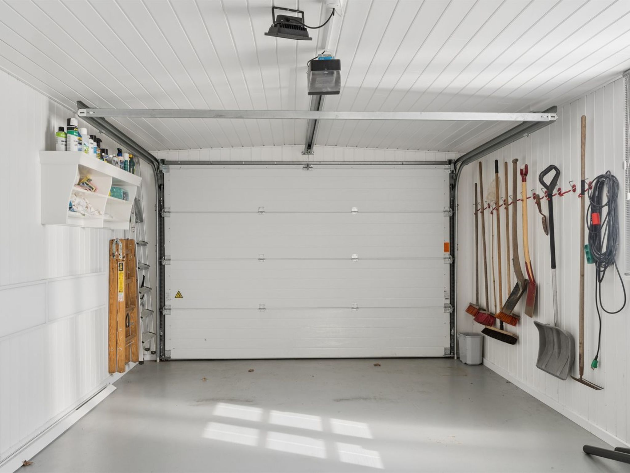 Eine Garage mit einer weißen Rolltür, Regalen voller Werkzeug und Ausrüstung sowie einem an der Decke befestigten Projektor.