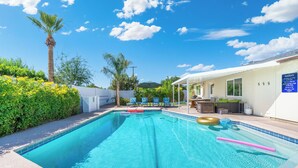 Una piscina con agua azul cristalina, rodeada de vegetación y un edificio blanco con techo azul.