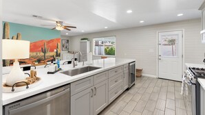Una cocina moderna con una isla grande, armarios blancos, ventilador de techo y ventana con vistas a la vegetación.
