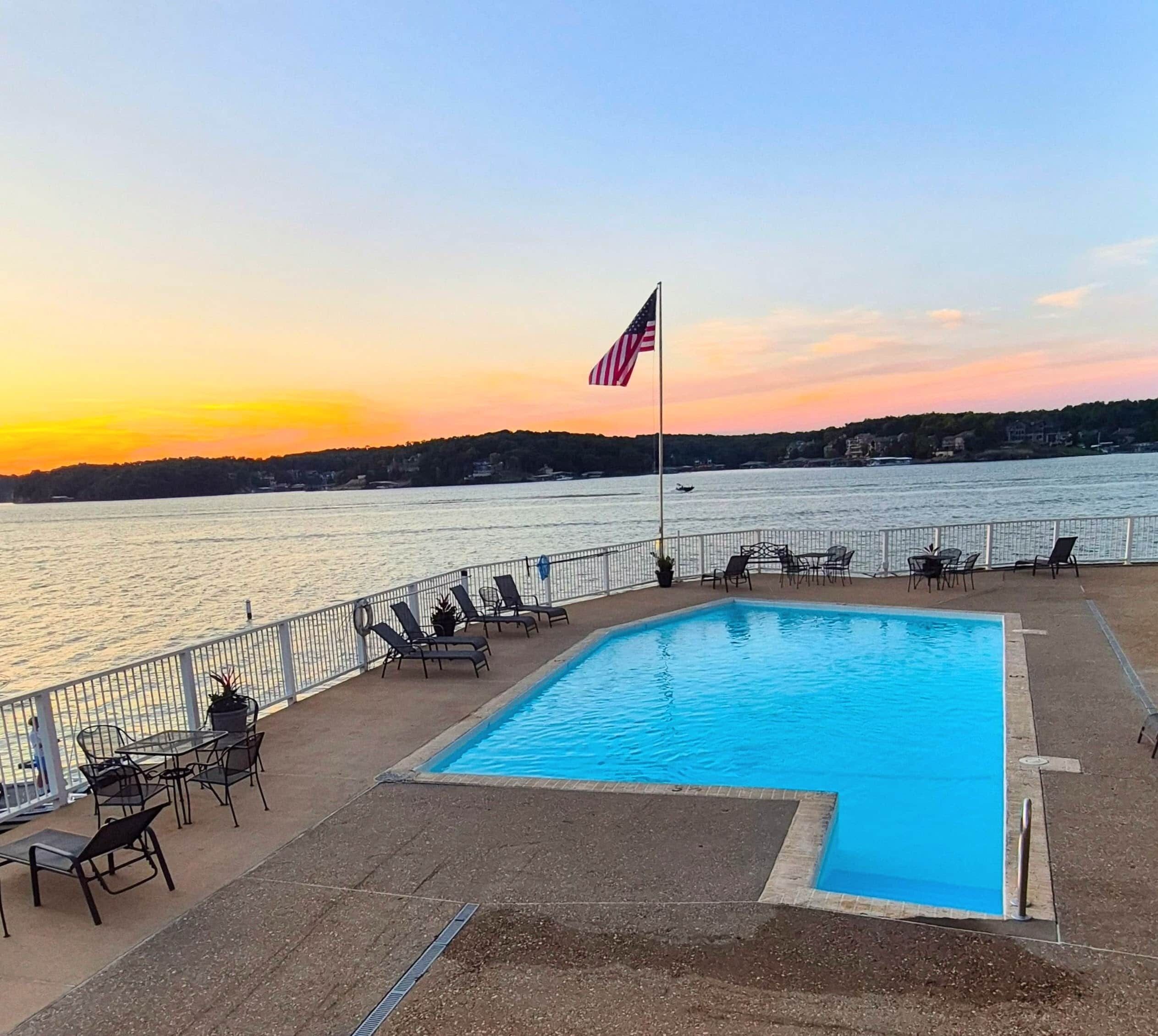 A swimming pool with lounge chairs and a flag, overlooking a body of water during sunset.