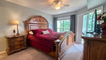 Une chambre à coucher avec un lit en bois, une table de chevet, une commode, un ventilateur de plafond et une fenêtre avec des rideaux.