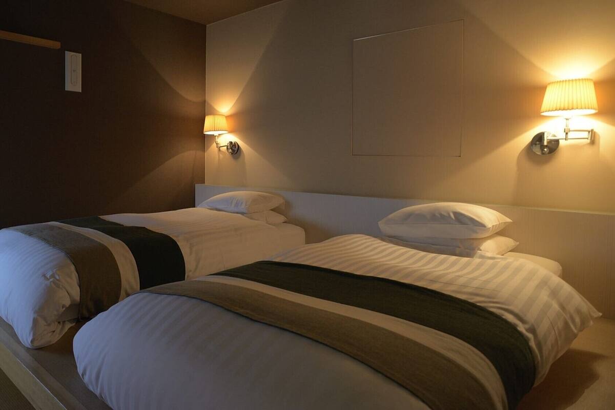 Habitación de hotel con dos camas, cada una con ropa blanca y mantas a rayas, dos lámparas fijadas a la pared y un cabecero de madera.