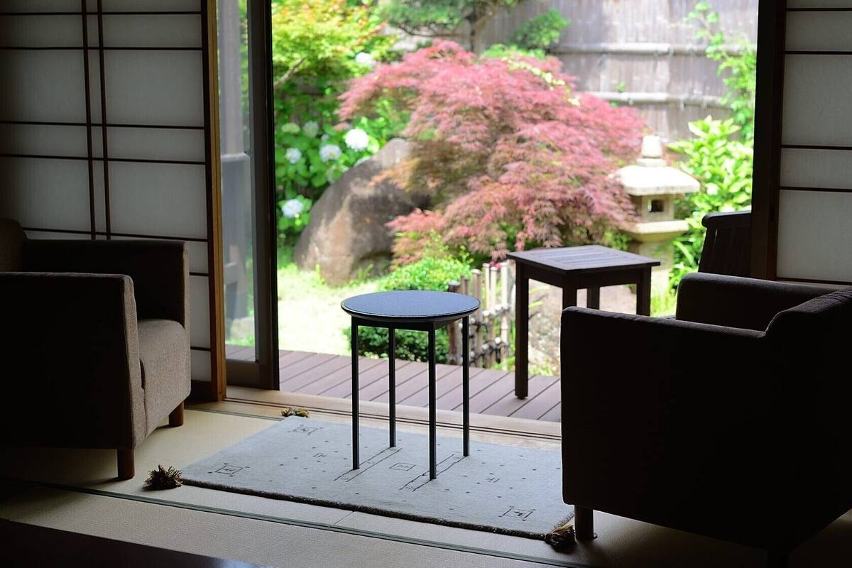 Una habitación con esteras de tatami, puertas correderas y vista a un jardín con un arbusto rojo.
