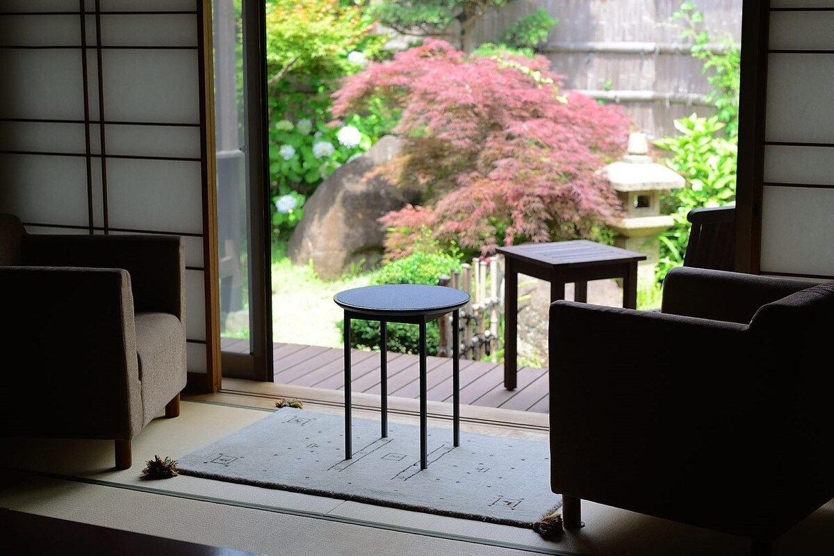 A room with tatami mats, a sliding door, and a view of a garden with a red bush.