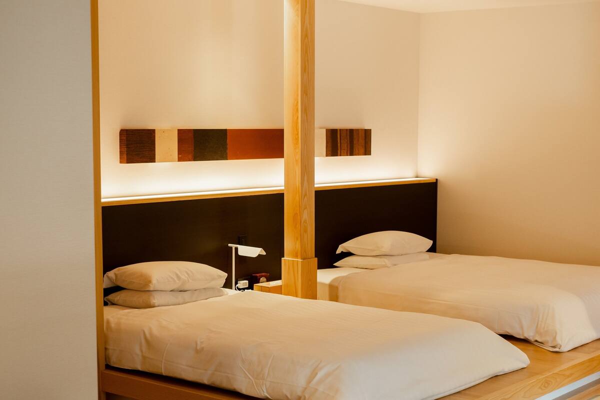 Habitación de hotel con dos camas individuales, cabecera de madera, mesita de noche con lámpara y una obra de arte en la pared.