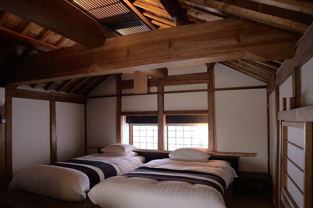 A traditional Japanese-style room with tatami flooring, a low ceiling with wooden beams, a sliding door, and a window with a view.