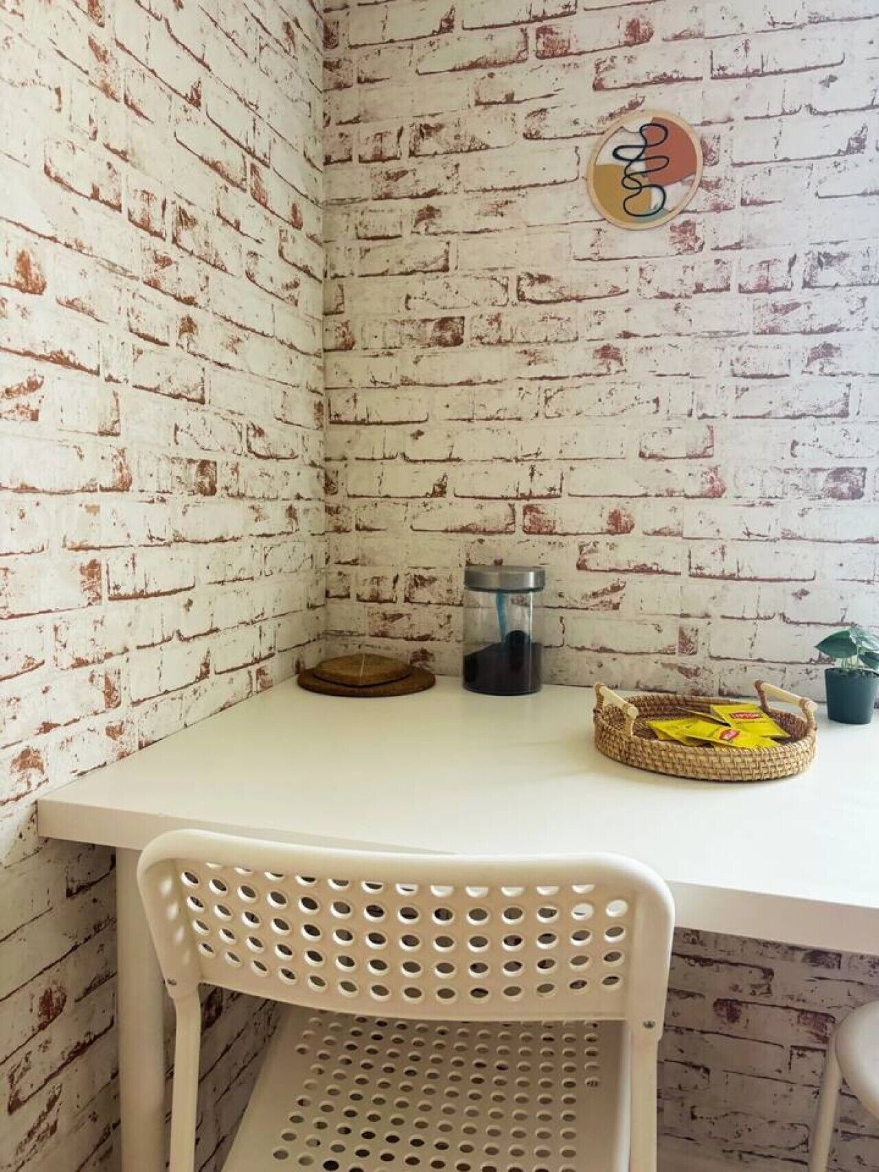 A corner of a room with a white table, a white plastic chair, a woven basket, a glass container, and a small potted plant.