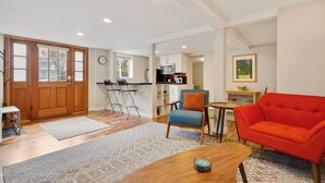 Una sala moderna con un sofá rojo, un sillón azul y una mesa de centro de madera. Hay una zona de cocina con un microondas y una mesa de comedor.