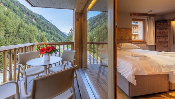 A room with a bed, a table with chairs, and a view of a forested mountain.
