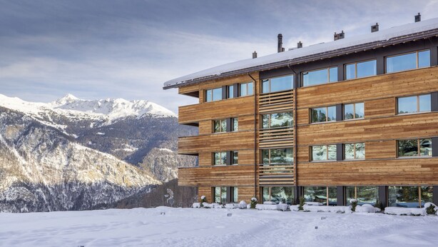 Un bâtiment en bois à plusieurs étages, doté de grandes fenêtres, se dressant sur fond de montagnes enneigées.