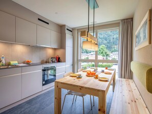 A modern kitchen with a dining area, featuring a large window with a view of a forested area.