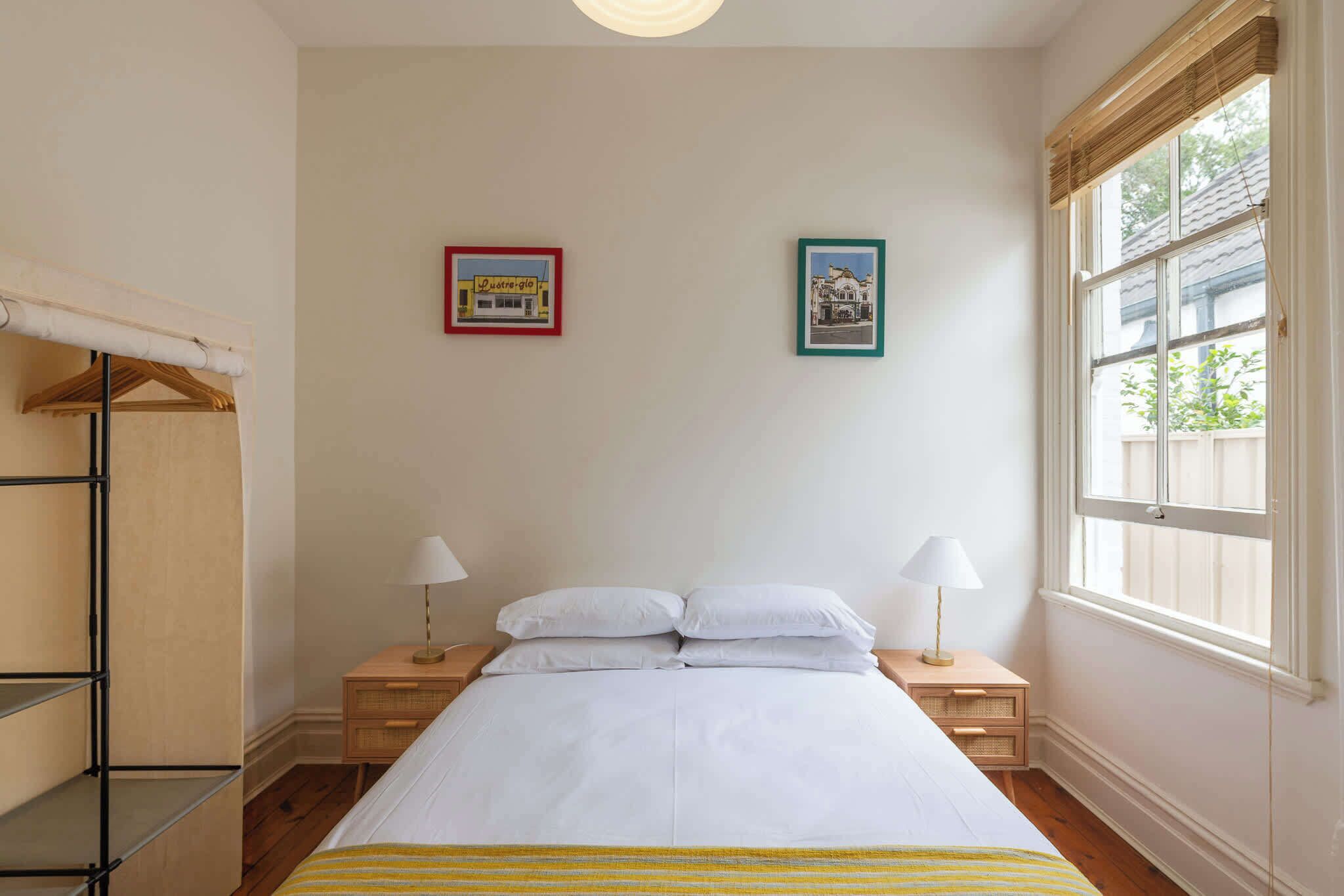 Un dormitorio con cama, dos mesitas de noche con lámparas, una ventana con persianas y dos cuadros en la pared.