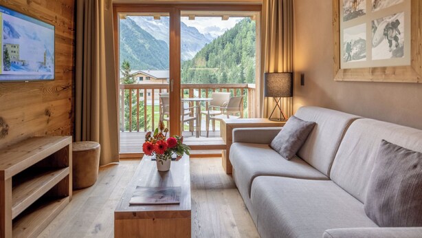 Una stanza con arredamento in legno, un divano, un tavolo con un vaso di fiori, una TV a parete e un balcone con vista sulle montagne.