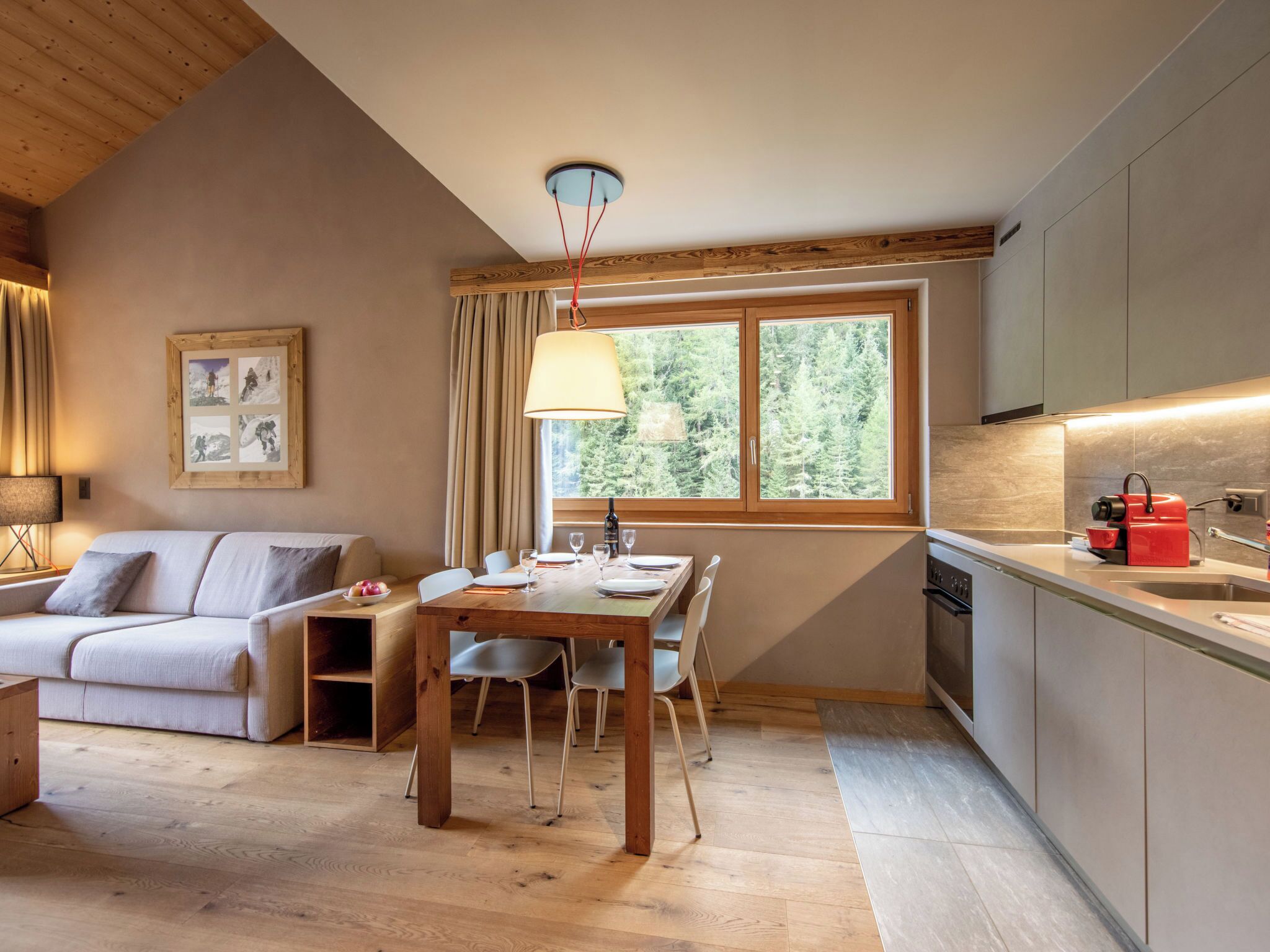 A cozy room with a wooden ceiling, a dining table with chairs, a sofa, a window with curtains, and a kitchen area with cabinets and a sink.