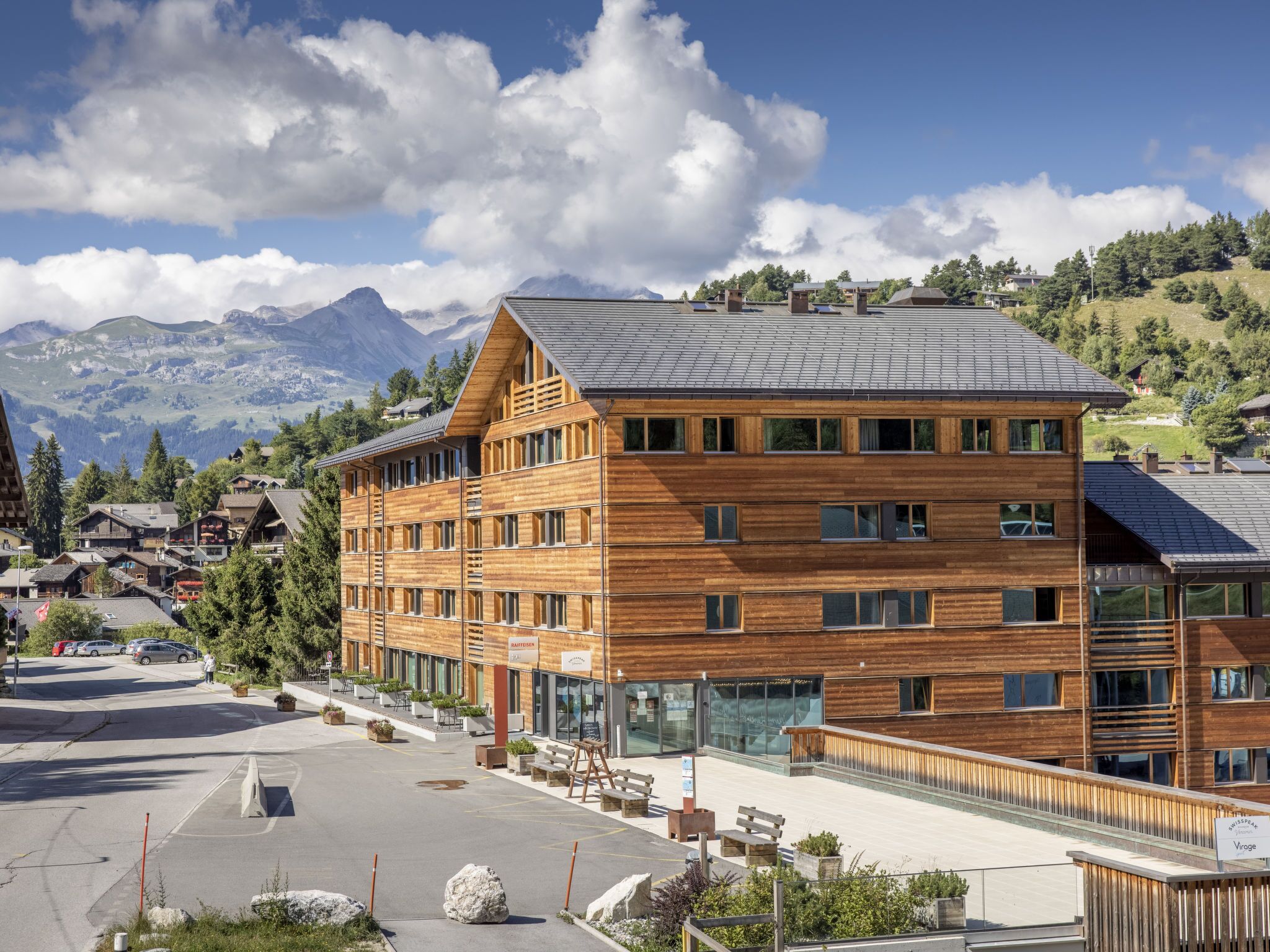 A modern wooden building with multiple floors, large windows, and a view of mountains in the background.