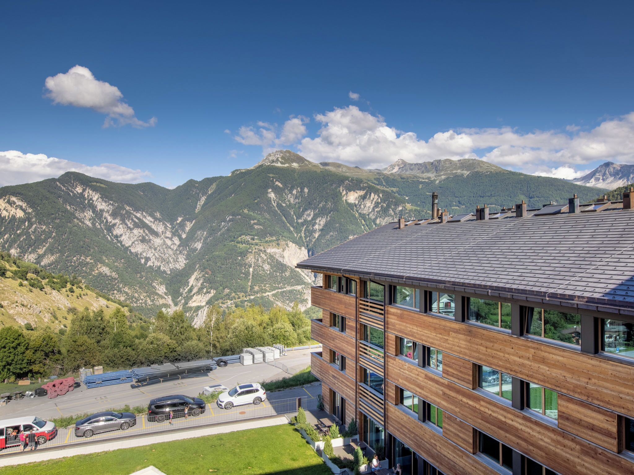 Un bâtiment moderne doté d’un toit en pente, de grandes fenêtres et d’un parking où sont garées des voitures.