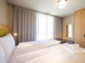 Une chambre d’hôtel avec deux lits, une grande fenêtre avec des rideaux, une tête de lit en bois, un miroir et un petit coin salon.