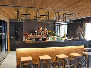 Een bar met houten plafond, barstoelen en een toonbank met een bartender.