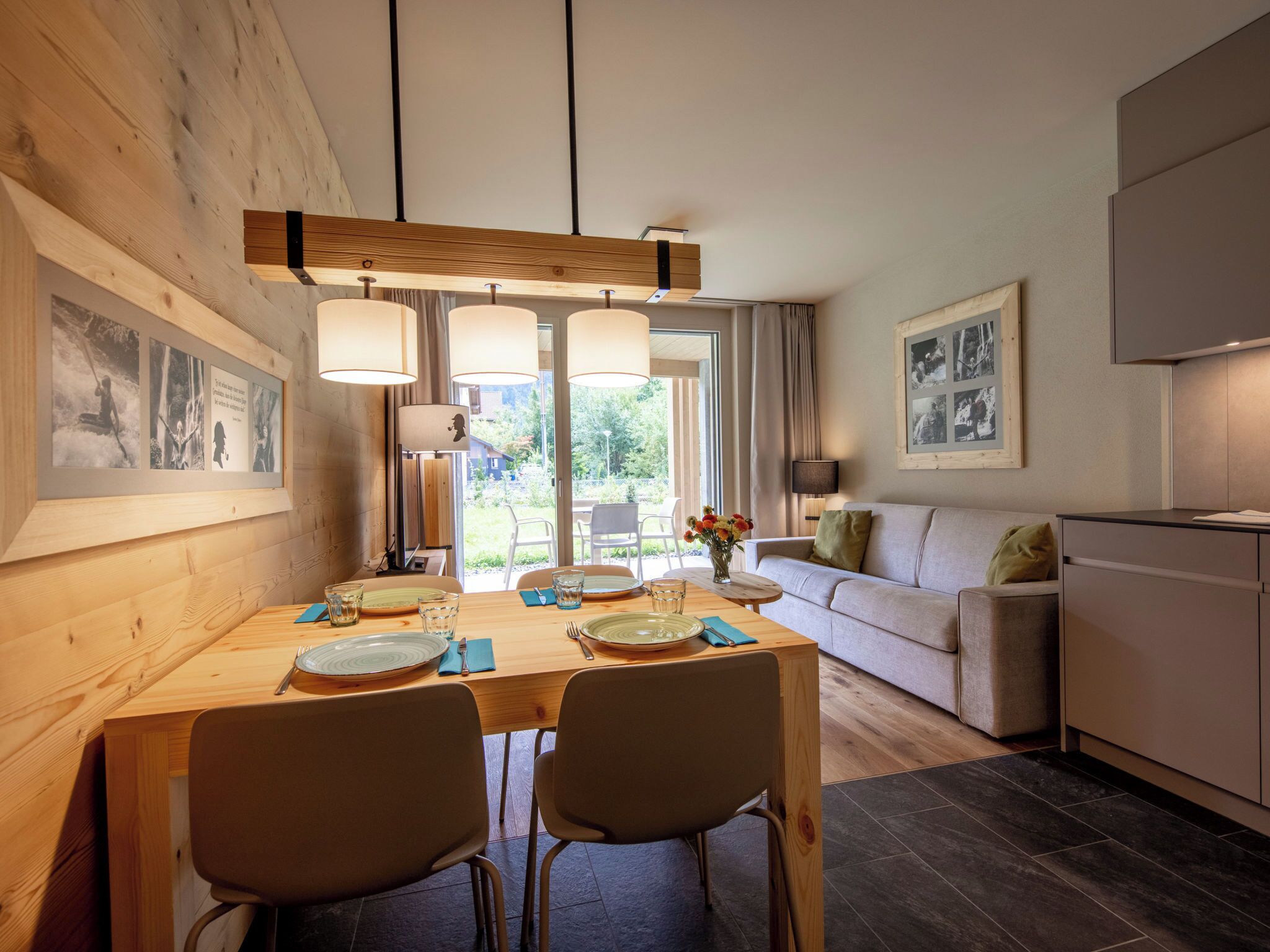 Un espace repas moderne avec une table en bois, des chaises et des luminaires suspendus. On aperçoit un canapé et un coin cuisine à l’arrière-plan.