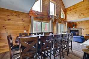 A dining area with a wooden table and chairs, a fireplace, and a view of the outdoors through large windows. - Poconos Chalet | Hot Tub & Game Room | Sleeps 16 (Blakeslee)
