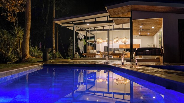 Ein modernes Haus mit einem wintergartenartigen Essbereich, einem Pool mit blauer Beleuchtung und einer Terrasse mit Gartenmöbeln.