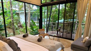 Ein Schlafzimmer mit einem großen Bett, einem Sessel und Blick auf einen üppigen Garten durch große Fenster.