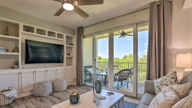A living room with a flat-screen TV, ottomans, a coffee table, a ceiling fan, and a sliding glass door leading to an outdoor patio. - Bahia Beach Calm Ruskin Retreat (Ruskin)