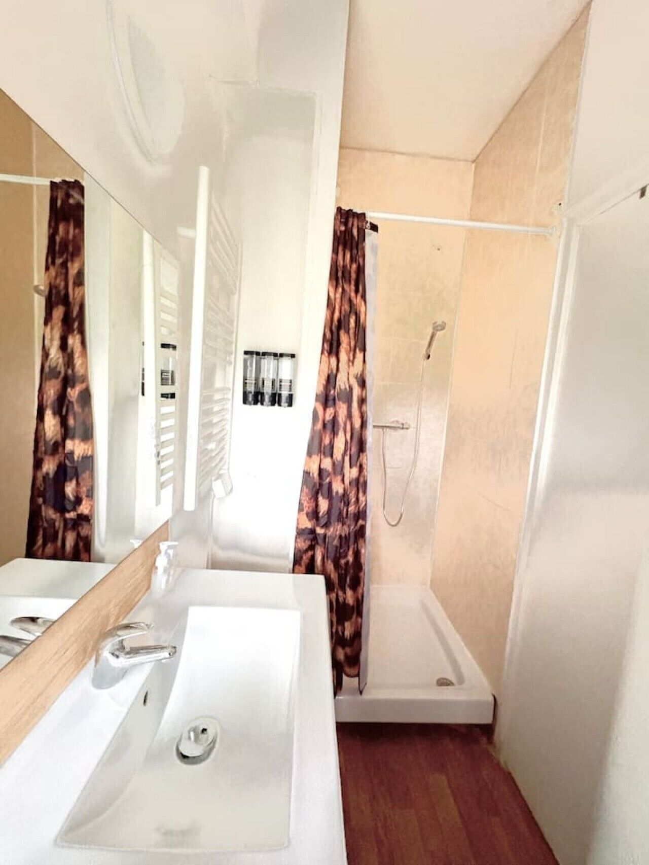 Une salle de bain avec un lavabo blanc, un miroir, une douche avec un mur beige et un rideau à motifs.