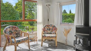 Quarto com uma grande janela, lareira, duas poltronas de vime com estofamento floral e uma pequena mesa com objetos decorativos.