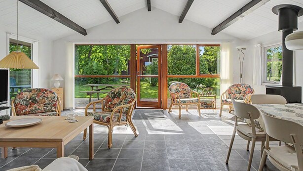 Um quarto espaçoso com teto alto, amplas janelas e vista para o exterior. Há várias cadeiras de vime e uma mesa de jantar com cadeiras.