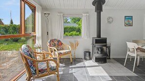 Quarto com uma grande janela, lareira, cadeiras de vime, uma mesa e vista para o verde.