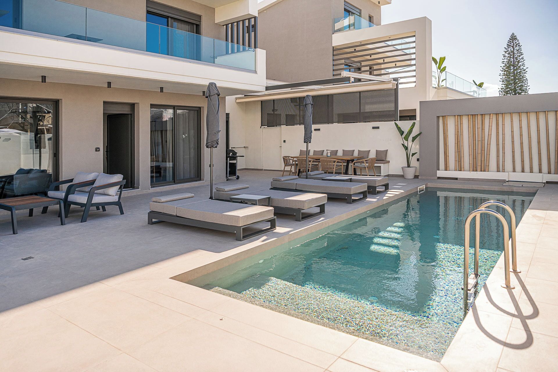 A modern outdoor pool area with lounge chairs and a table, surrounded by a building with large windows and a balcony.