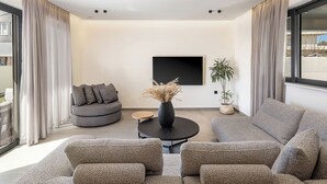 A modern living room with a sectional sofa, a round coffee table, and a comfortable armchair. A large black artwork hangs on the wall, and there is a potted plant on the floor. - The Ekfrasis- Mossa Concept Villas (Chania)
