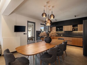 A modern kitchen with dark cabinetry, a marble countertop, and a dining area with a wooden table and patterned chairs. - The Ekfrasis- Mossa Concept Villas (Chania)