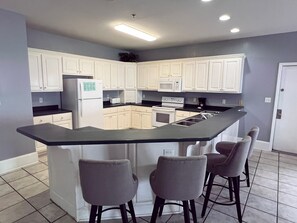 A kitchen with white cabinets, a black countertop, a built-in island with bar stools, and a microwave. - Sea Oats Motel (Destin)