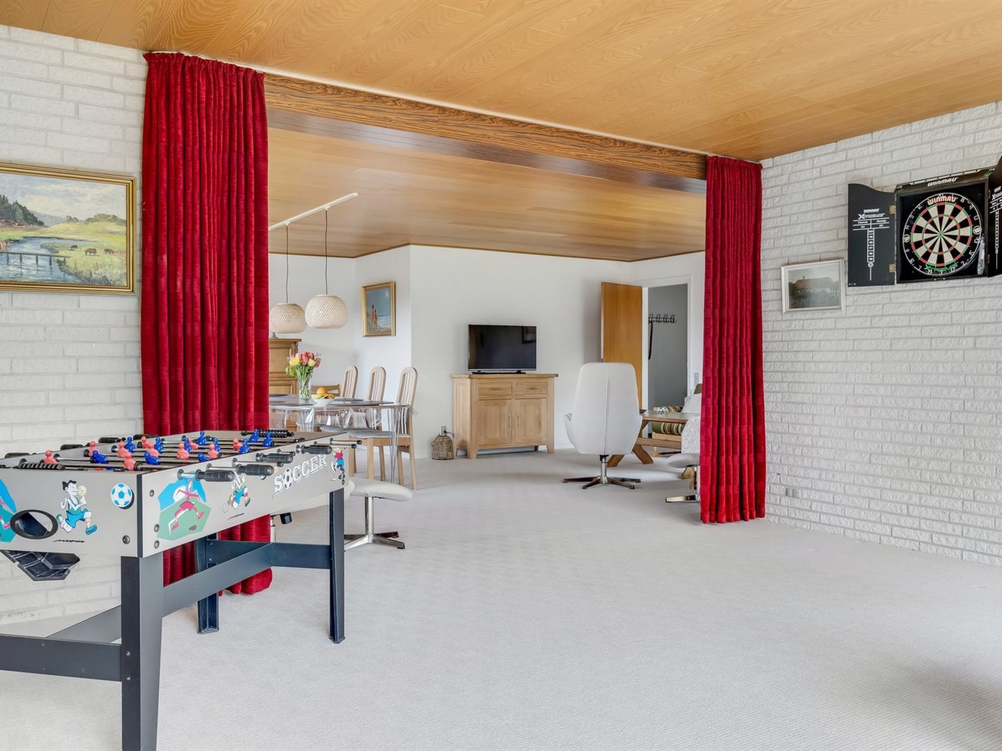 A room with a foosball table, a dartboard, a television, and a dining area with chairs and a table.