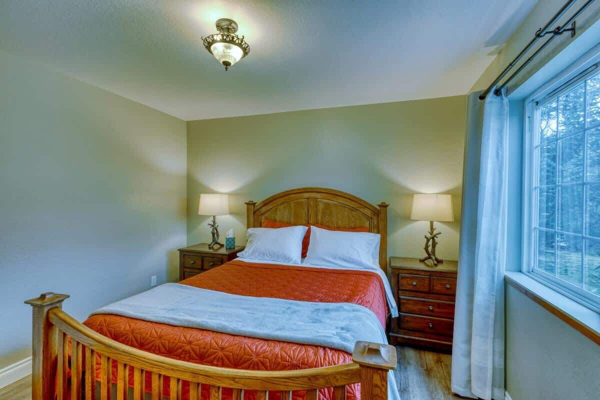A bedroom with a wooden bed, two bedside tables, a nightstand, a window with curtains, and a ceiling light fixture.
