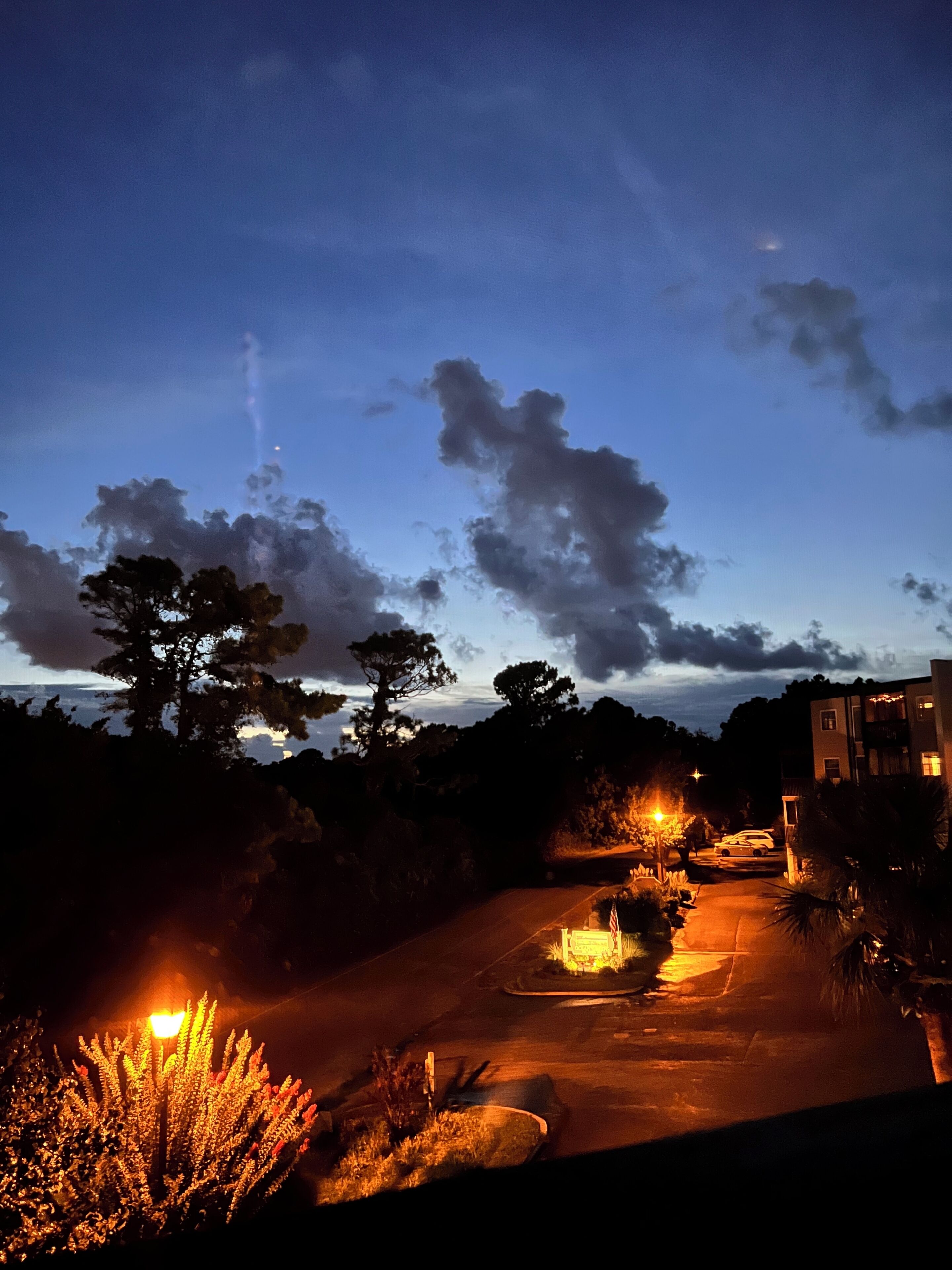 A nighttime view of a parking area with a building in the background, trees, and a street lamp.