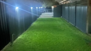 A nighttime view of a well-lit pathway leading to a building with a metal fence and trees in the background.