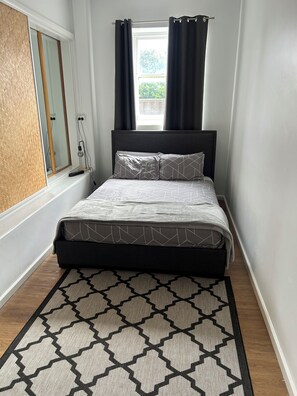 Une chambre à coucher avec un lit, une fenêtre avec des rideaux, une table de chevet et un tapis à motifs.