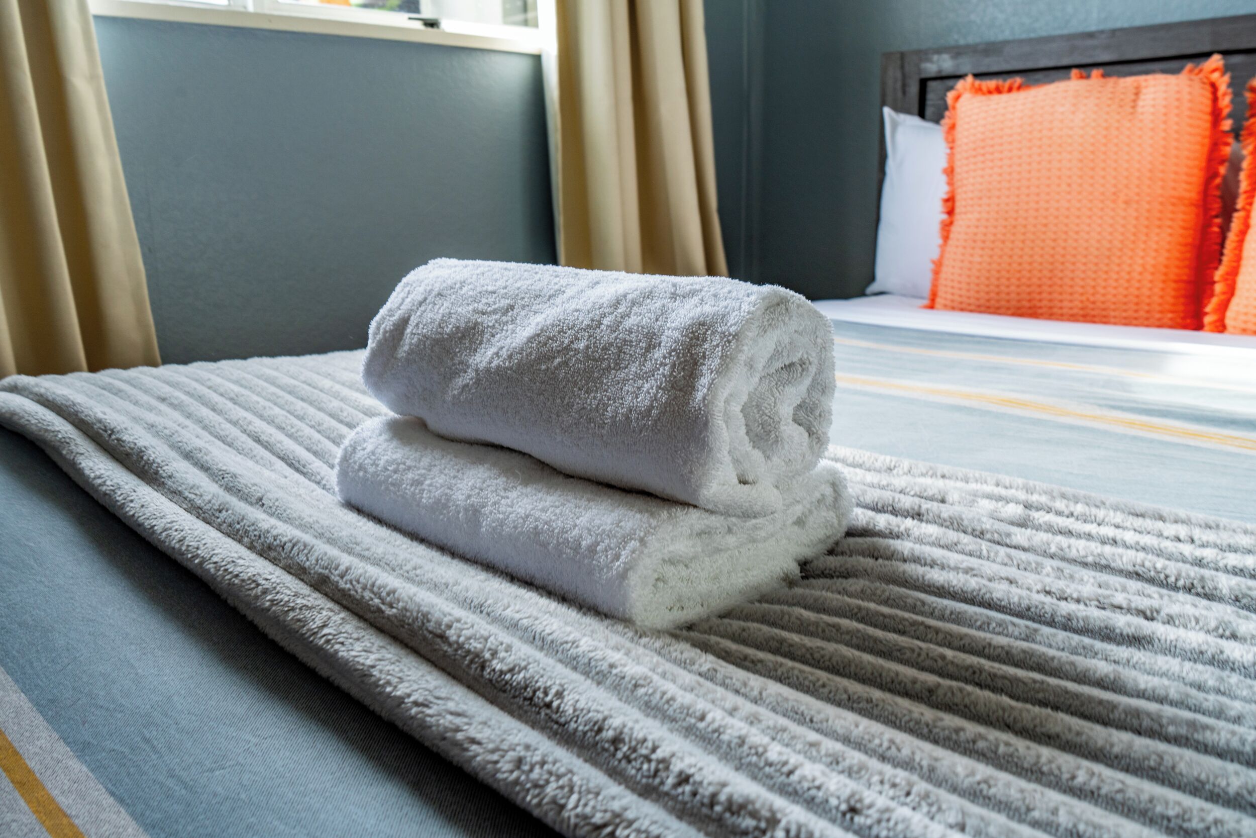 A neatly made bed with a folded white towel, a striped blanket, and an orange pillow.