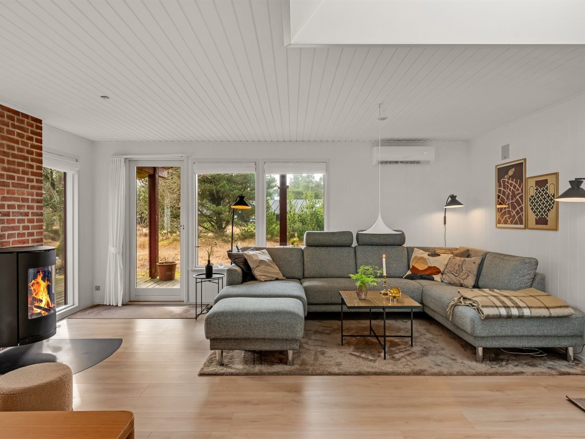 Un salon moderne avec une cheminée, un canapé d’angle, une table basse et une grande fenêtre donnant sur la forêt.