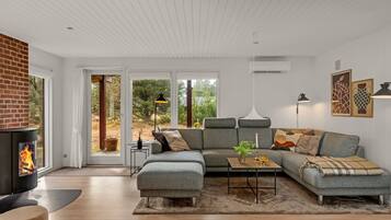 Un salon moderne avec une cheminée, un canapé d’angle, une table basse et une grande fenêtre donnant sur la forêt.