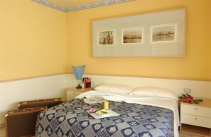 Habitación de hotel con cama, mesita de noche, lámpara, un cuadro en la pared y una pequeña mesa con una flor.