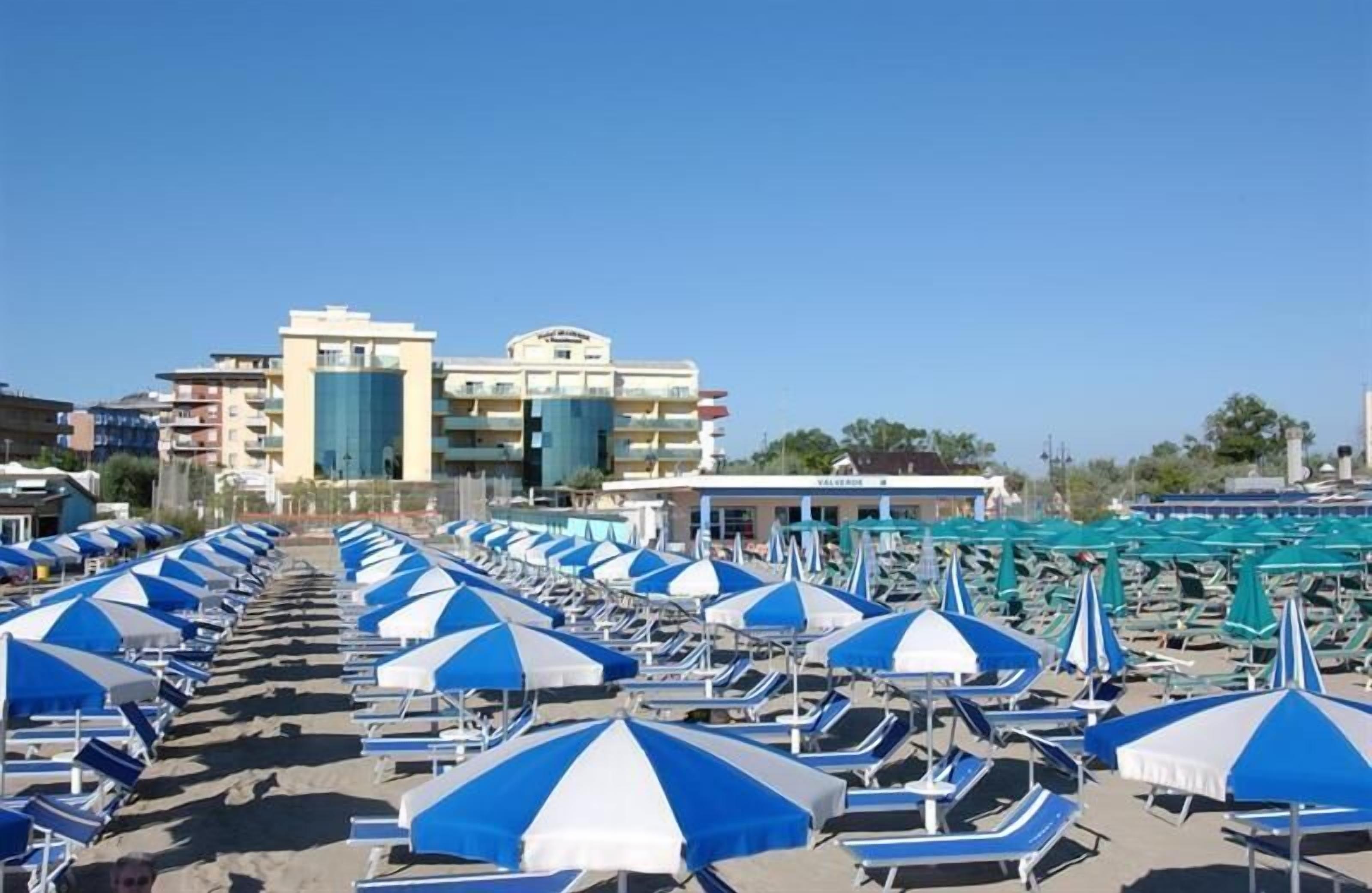 A beachside hotel with rows of sunbeds under blue and white umbrellas, a pool area, and a building with balconies.