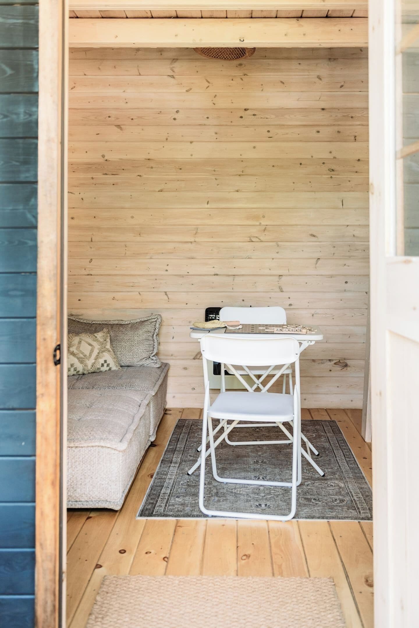 A small room with a wooden floor, a white folding table, a white chair, a sofa with a cushion, and a patterned rug.