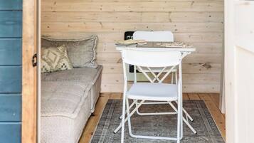 A small room with a wooden floor, a white folding table, a white chair, a sofa with a cushion, and a patterned rug.