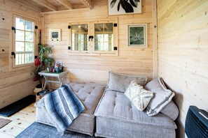 A cozy wooden cabin interior with a sofa, pillows, a small table with a plant, and framed pictures on the wall. - Off-Grid Getaway | Fireplace, BBQ & Patio (Gravenhurst)