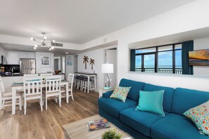 Ein modernes Wohnzimmer mit einem türkisfarbenen Sofa, einem Essbereich mit weißer Möbel und einer Küche mit weißen Schränken sowie einem großen Fenster mit Blick.